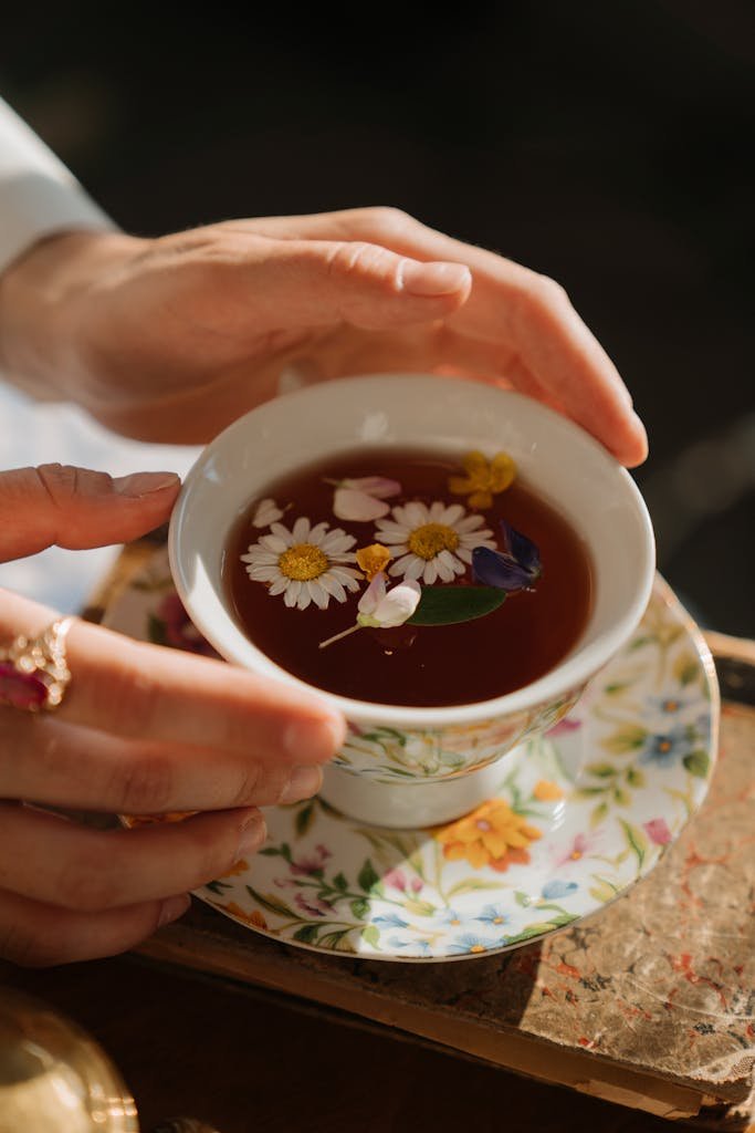 A woman holding a porcelain cup of chamomile tea with floating flowers in a delicate setting.